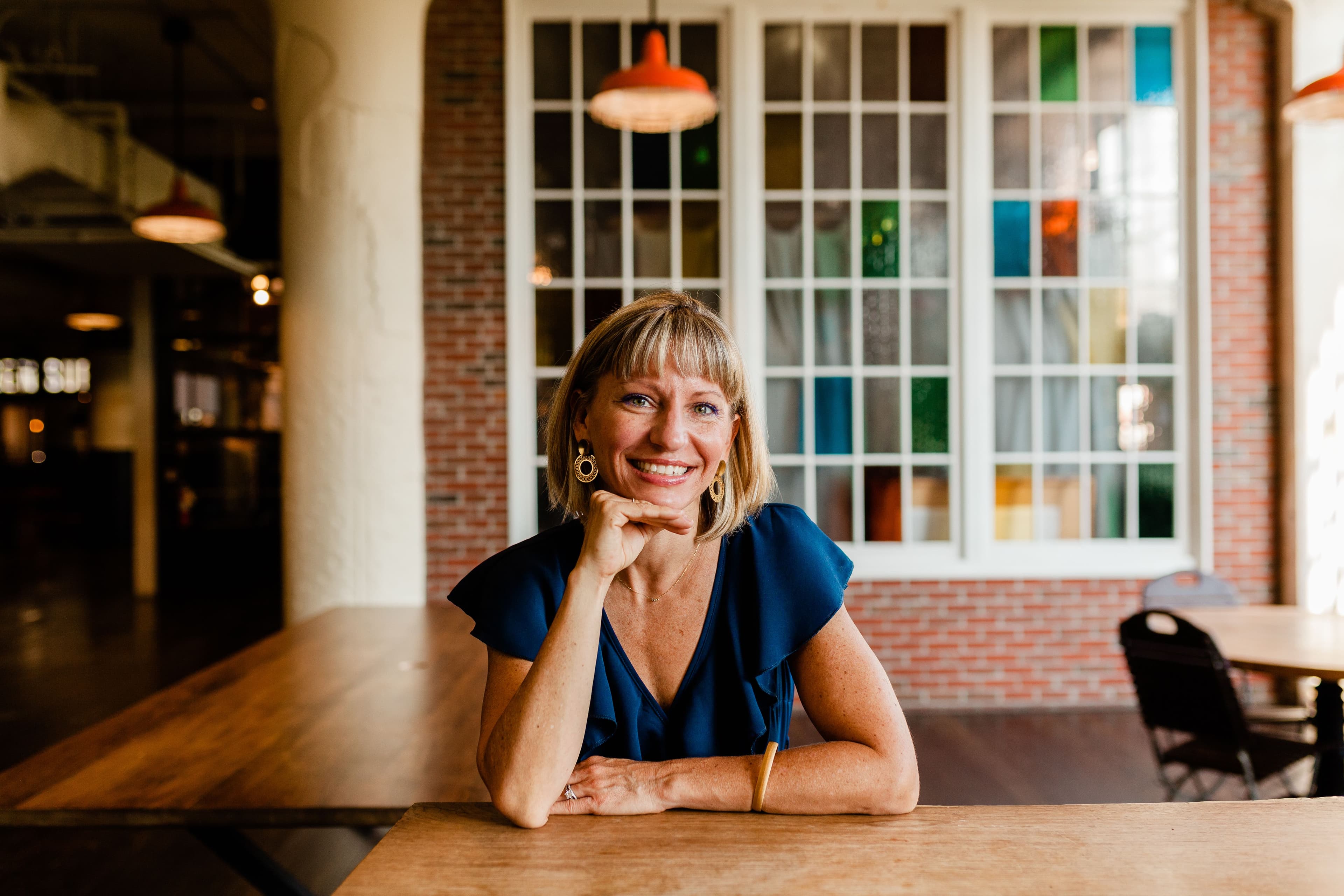 Blonde woman in blue top smiles, resting chin on hand at a wooden table.