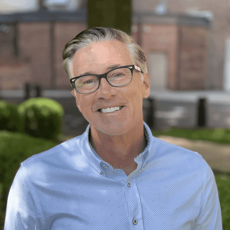 Smiling man with glasses and grey hair wearing a light blue shirt outdoors.