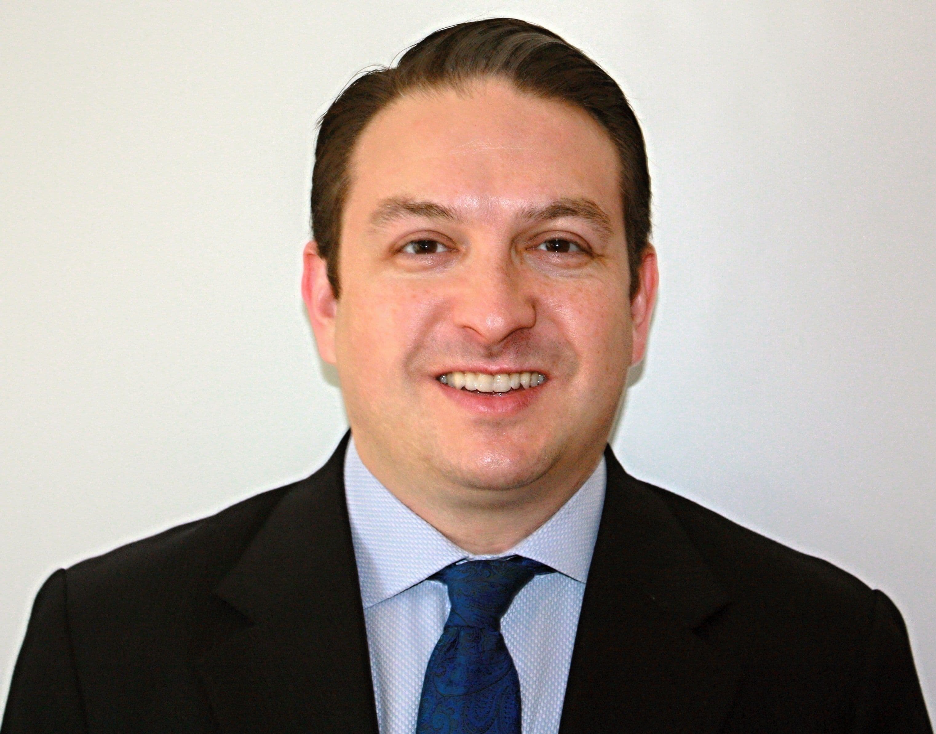 A smiling man in a dark suit and blue tie poses for a headshot.