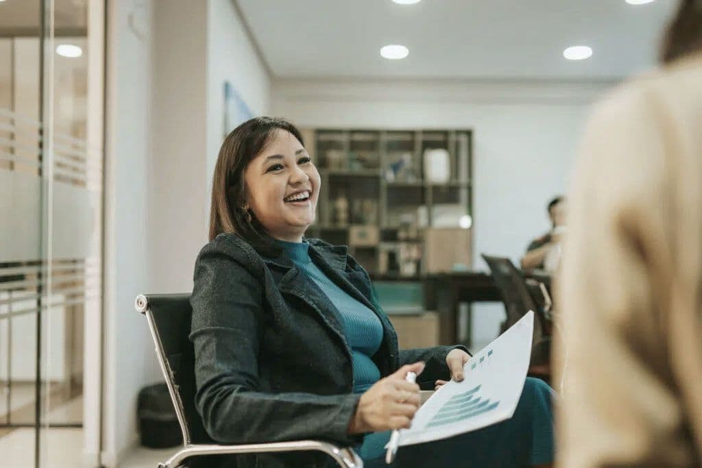 A woman in a suit laughing with some papers in her hand sitting at a conference table.