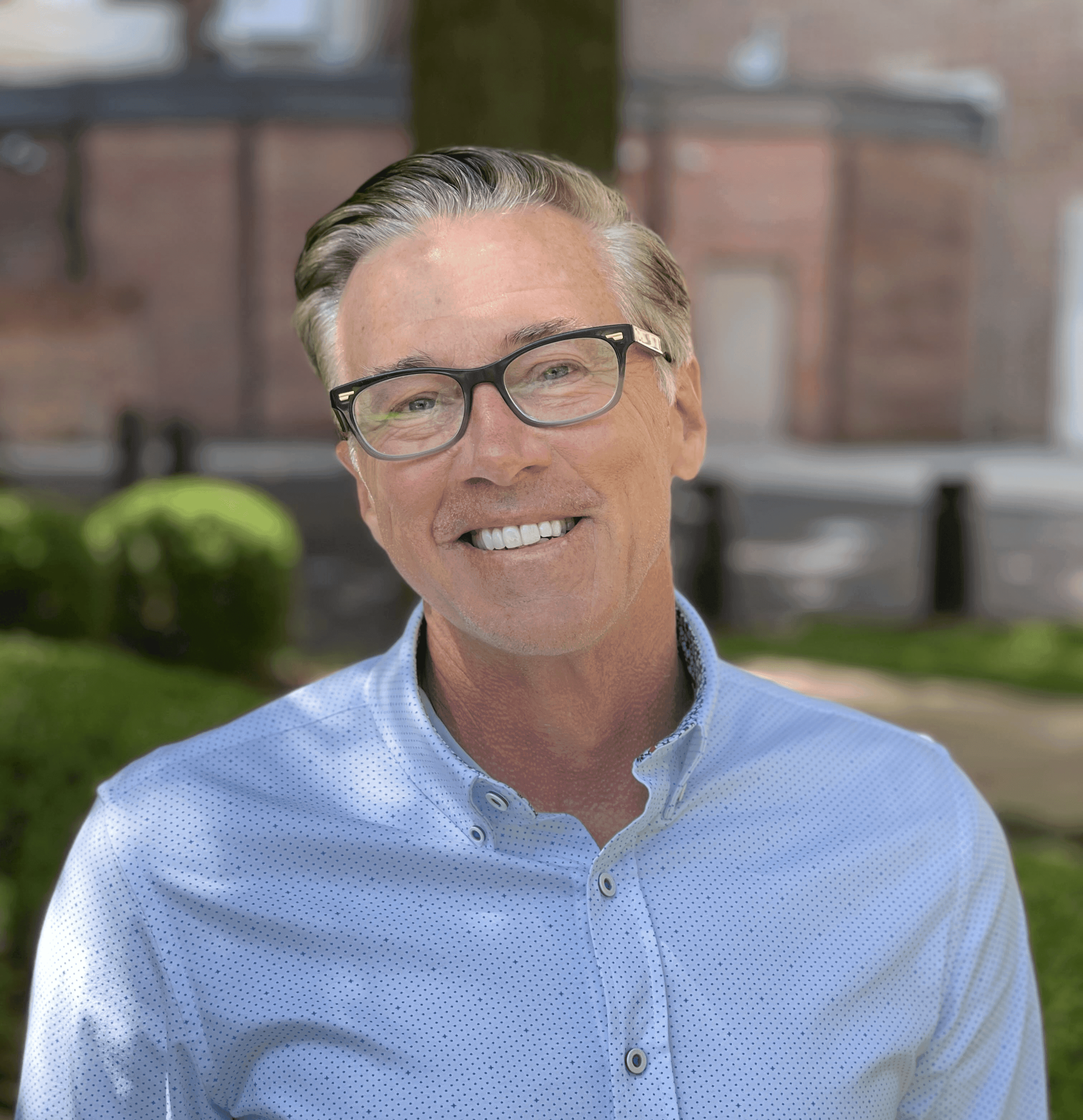 A middle-aged man with glasses smiles directly at the camera outdoors.