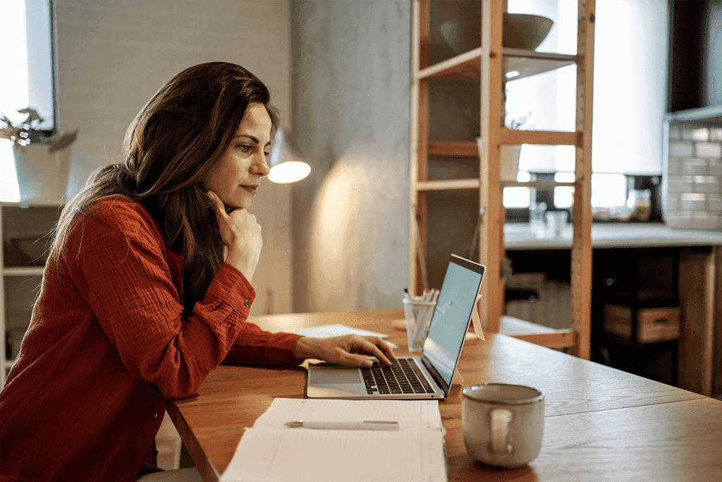 Woman in red shirt focused on laptop screen at a wooden desk.