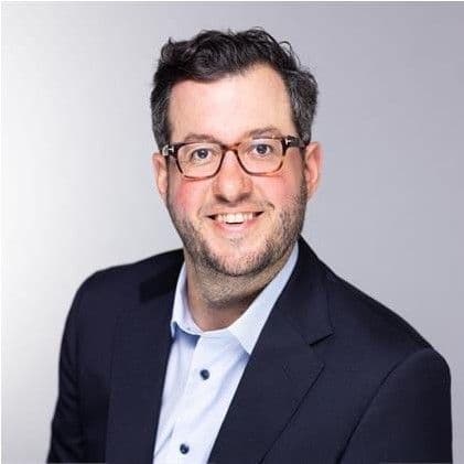 A smiling man with glasses and a beard wears a dark suit and a light blue collared shirt.