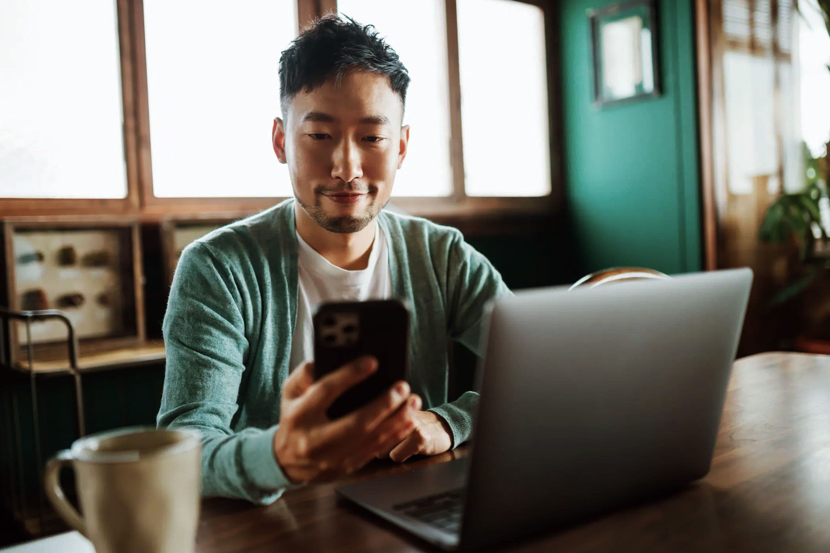 A man sitting at a coffee shop with a laptop on the table scrolling through his phone.